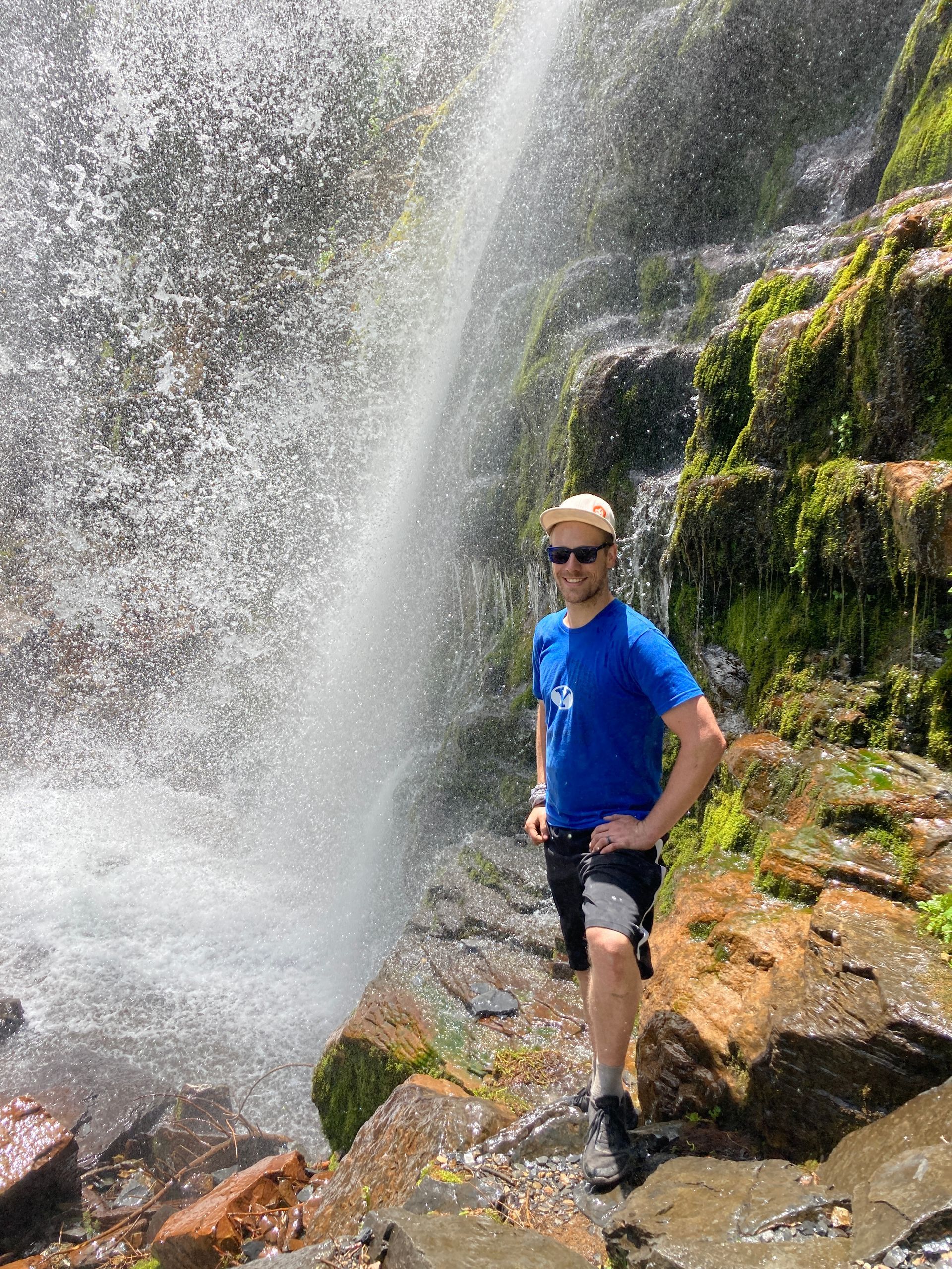 A man in a blue shirt is standing in front of a waterfall.