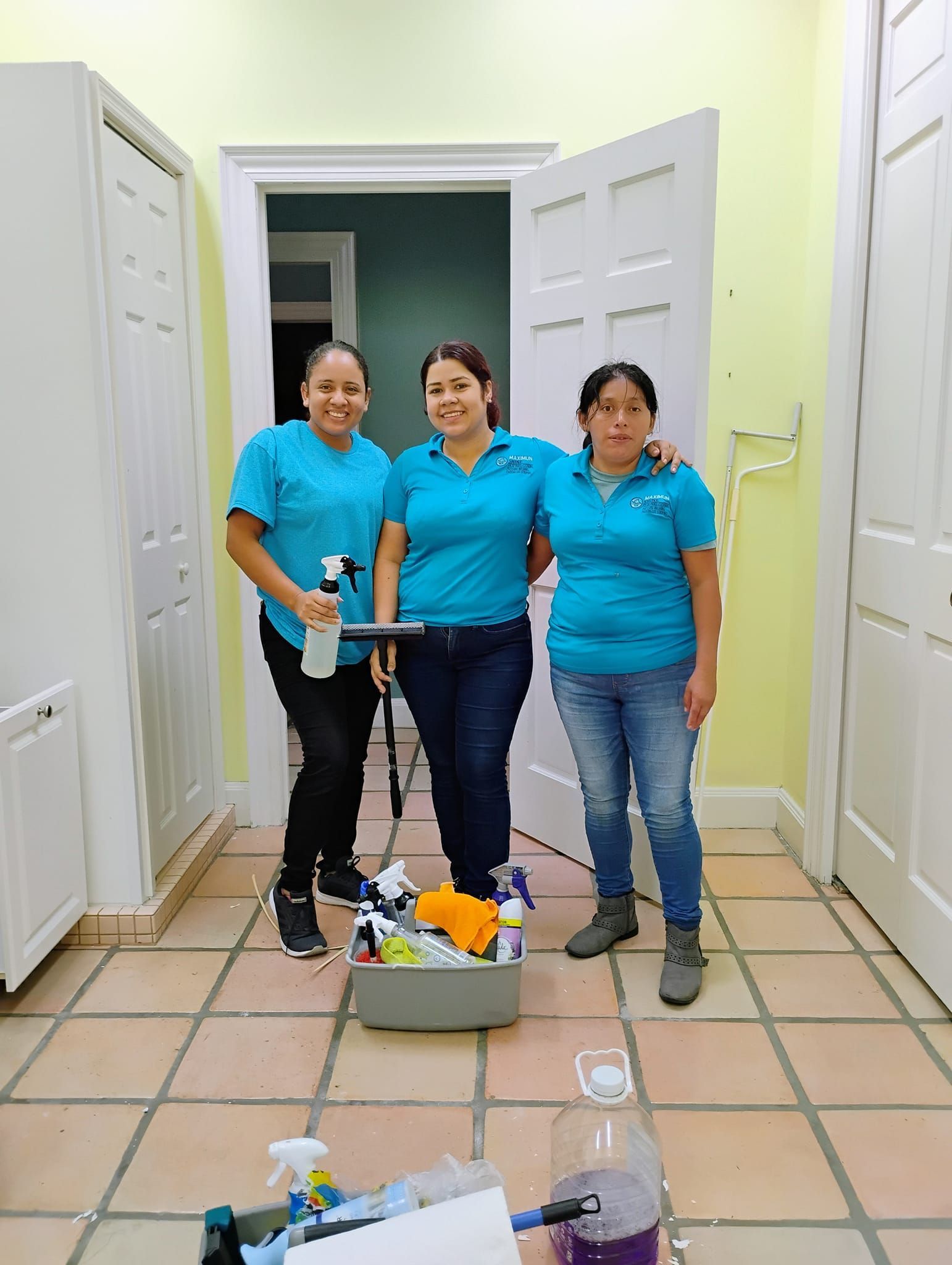 Three women are standing next to each other in a hallway holding cleaning supplies.
