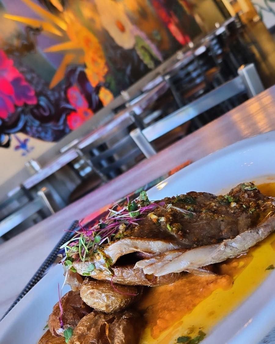 Plate of food: fish, potatoes, orange sauce, garnished with microgreens. Restaurant setting with artwork.