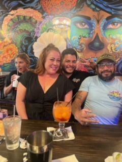 Four people at a bar, smiling. Colorful mural in background. Drinks on the counter.