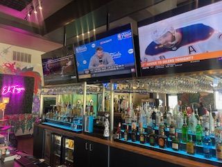 Bar with liquor bottles, glasses, and TVs showing sports; pink neon sign on left.