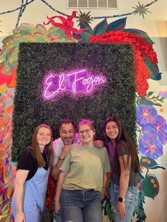 Group of four people smiling in front of a neon sign that says 