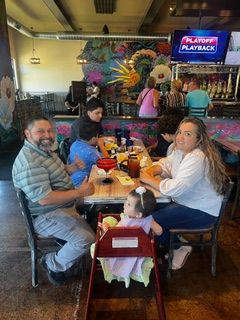 Family dining at a restaurant. A father smiles, a baby sits in a high chair, a mother eats, and others dine nearby.