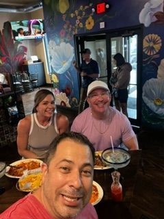 People at a restaurant, taking a selfie. Smiling man in foreground, others eating. Colorful floral wall.
