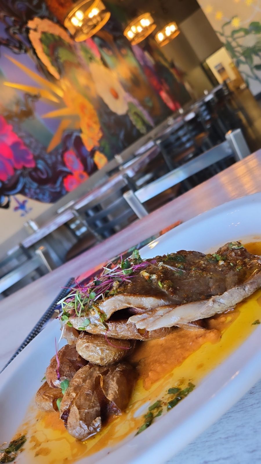 Grilled fish and roasted potatoes on a plate, in a restaurant with colorful wall art.