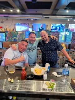 Three men smile at a bar. One gives a thumbs up, others have arms around each other. Chips, salsa, drinks, and TVs in background.