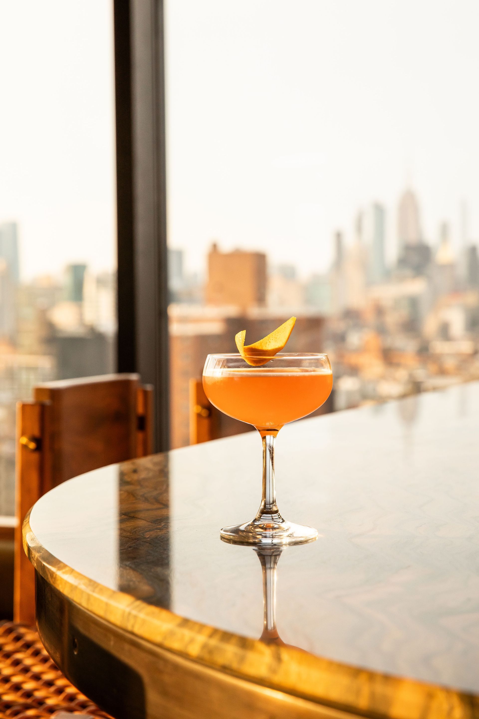 Cocktail in a coupe glass with a citrus peel garnish, on a reflective table overlooking a cityscape.