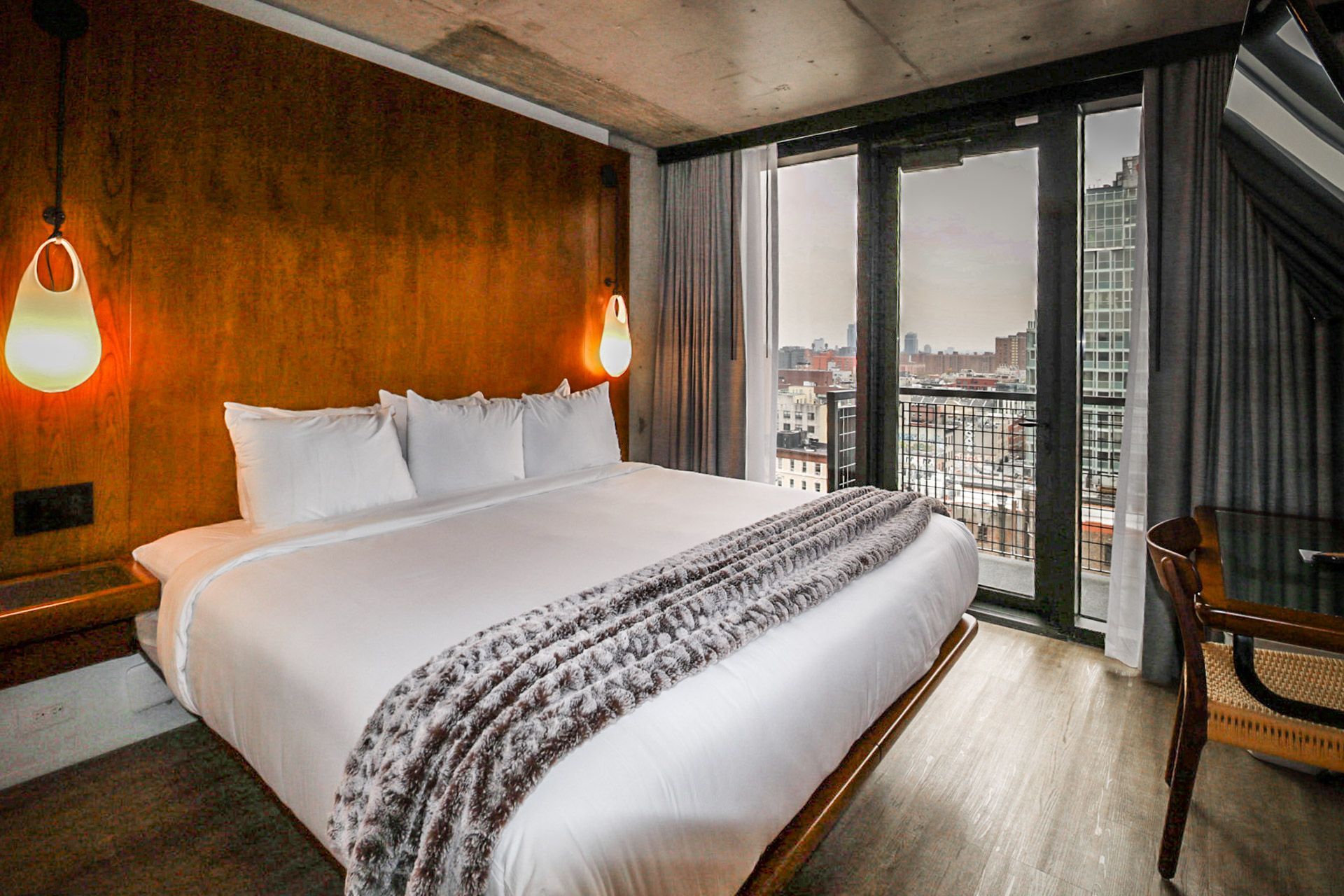 Hotel bedroom with large bed, wooden headboard, city view, and two hanging lights.