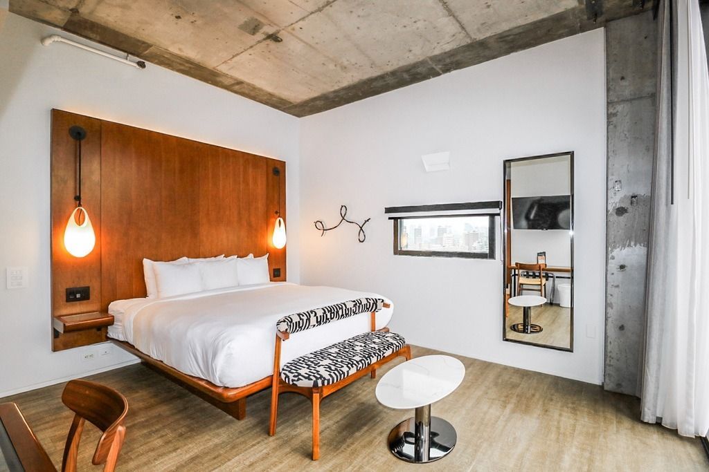 Hotel room with a bed, bench, chair, mirror, and small table. White walls, wood accents, and concrete ceiling.
