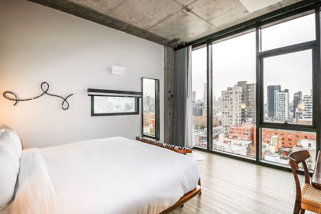 Bedroom with large windows overlooking a city, white bedding, exposed concrete ceiling, and decorative wall art.
