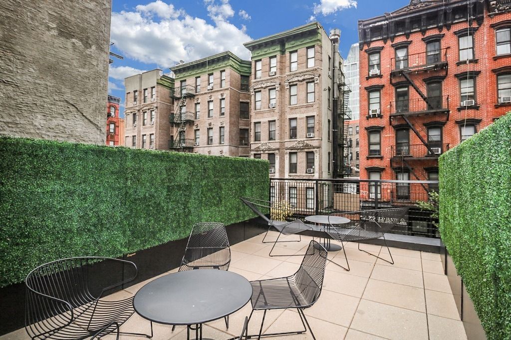 Rooftop patio with modern furniture; green hedges line the perimeter; multi-story buildings.