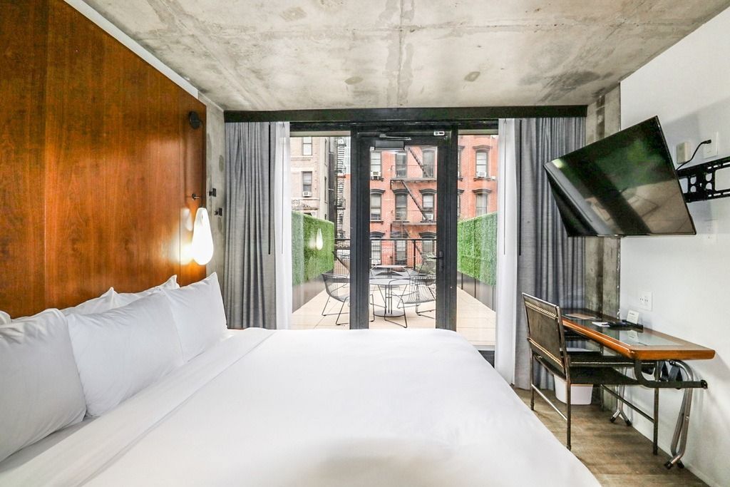 Hotel room with bed, balcony overlooking outdoor seating, and wall-mounted TV.