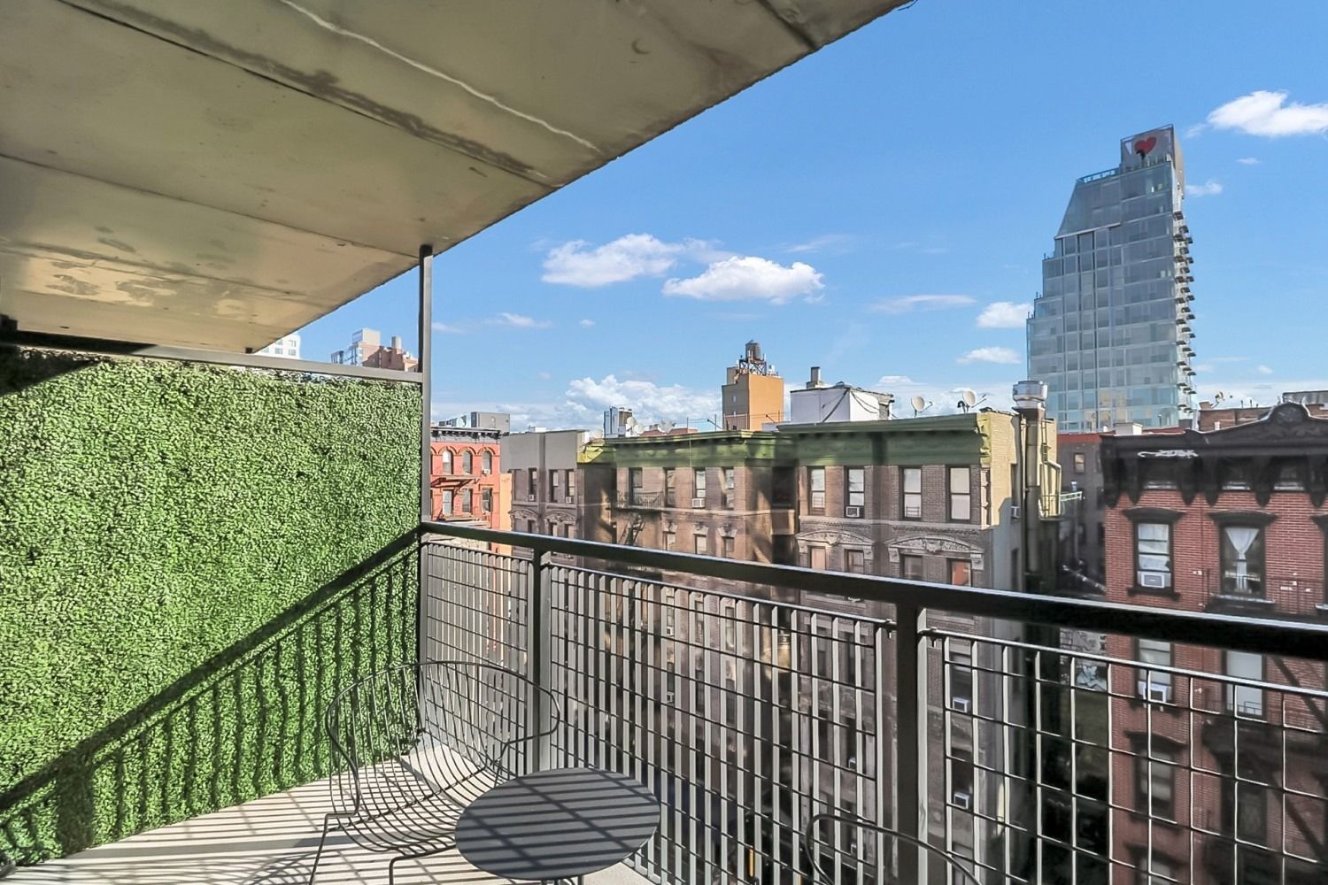 Balcony view of city buildings with blue sky. Green hedge on left, metal railing, small table.