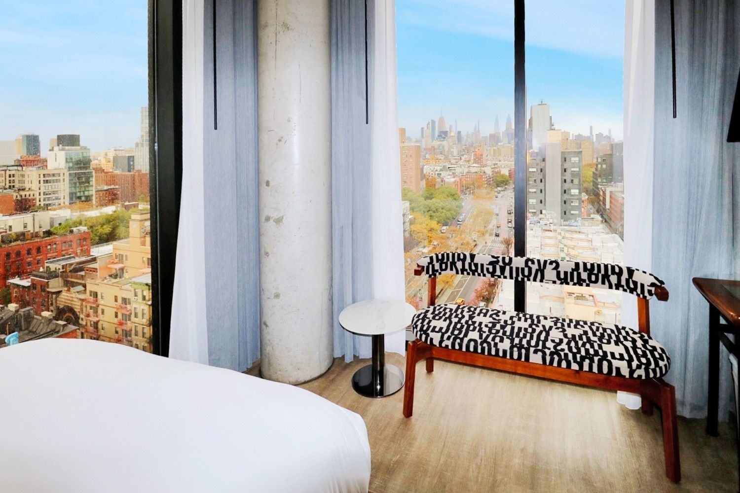 Bedroom interior with city view, bed, bench, side table, large window, and light blue sky.