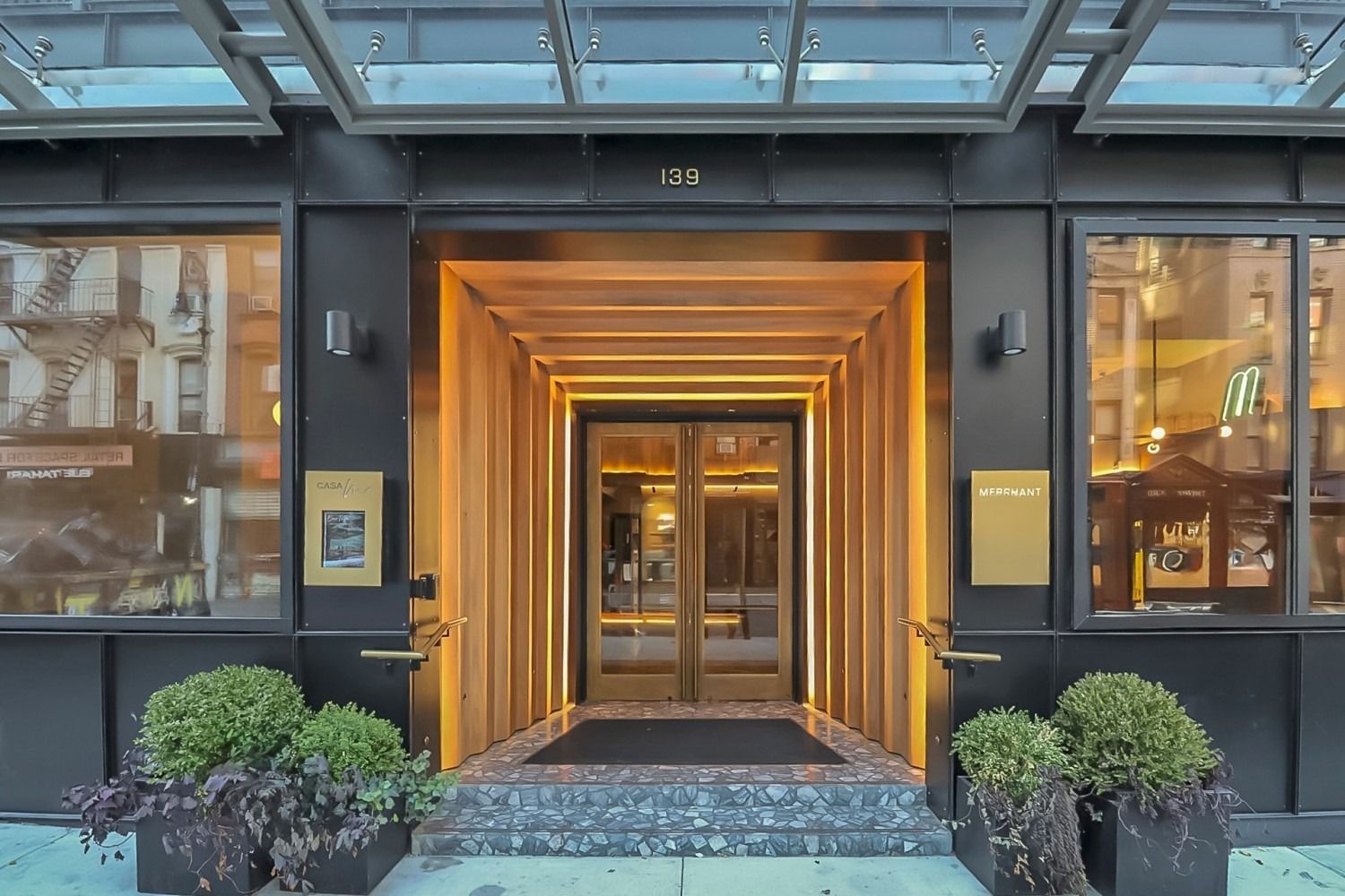 Hotel entrance with gold-lit wooden doorway, glass awning, and potted greenery.