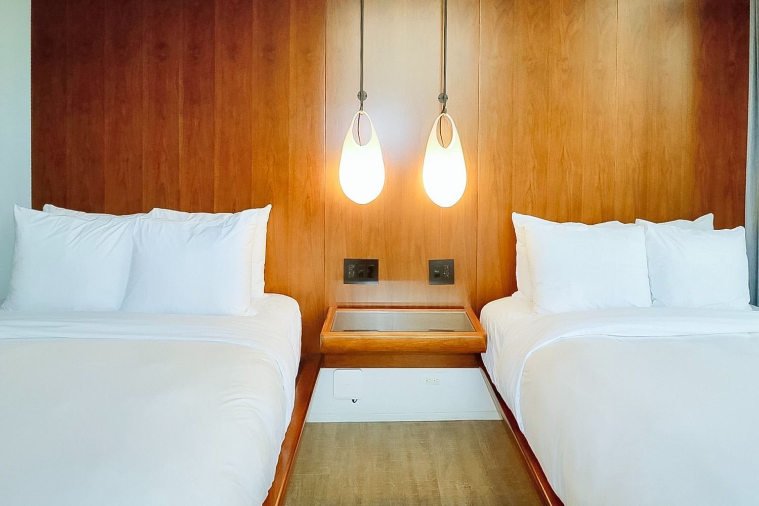 Two beds in a hotel room with white linens, wooden headboard, pendant lights, and nightstand.