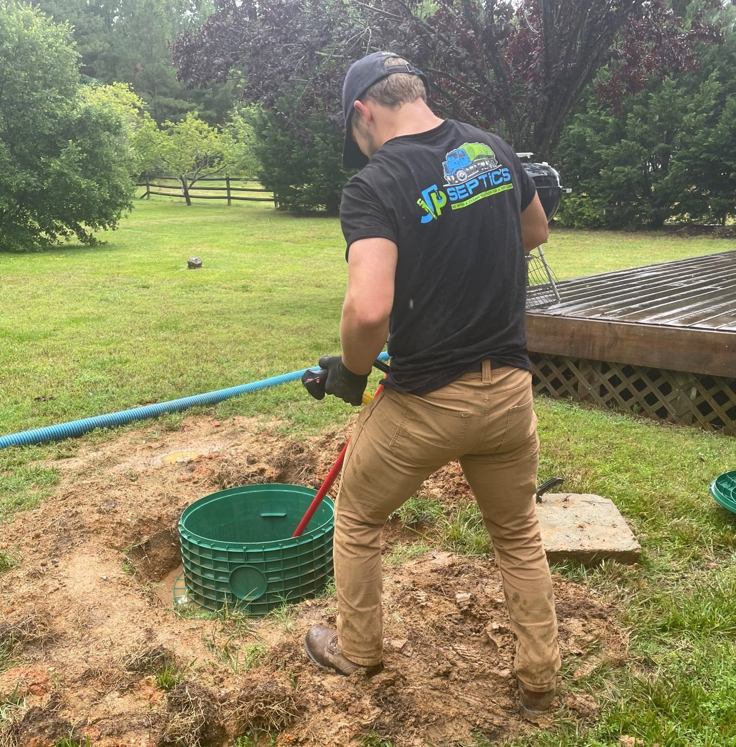 A man pumping a septic tank in a yard; he wears a black t-shirt, khaki pants, and a hat.