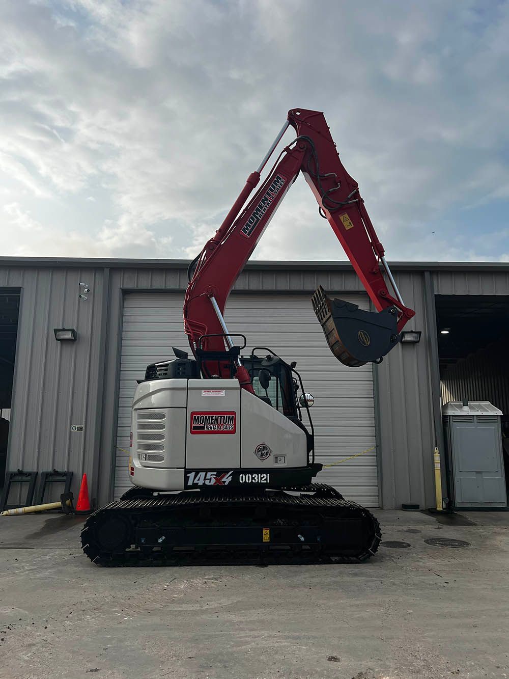 A red and white excavator is parked in front of a garage door.