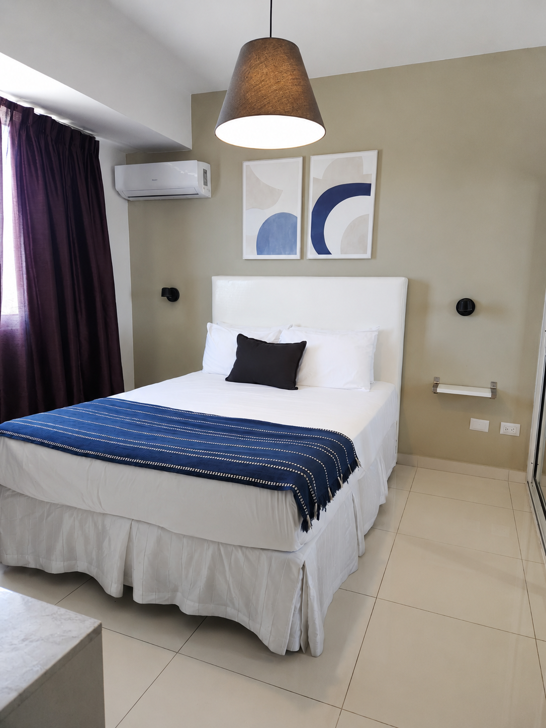 Habitación minimalista con cama, televisor sobre mueble blanco y gran ventanal con vistas a la ciudad.