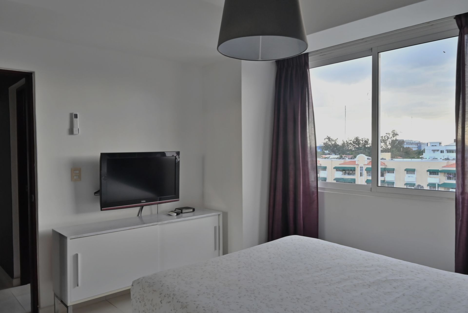 Habitación luminosa con cama, televisor sobre un mueble blanco y una gran ventana con cortinas con vistas a los edificios.
