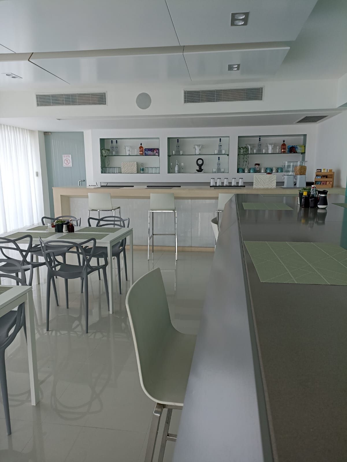 Cafetería luminosa y moderna con mesas, sillas y un mostrador de servicio detrás de estantes de cristal.