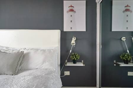 Dormitorio moderno con cama blanca, paredes grises, cuadro de un faro, armario con espejo y pequeña lámpara de estante.