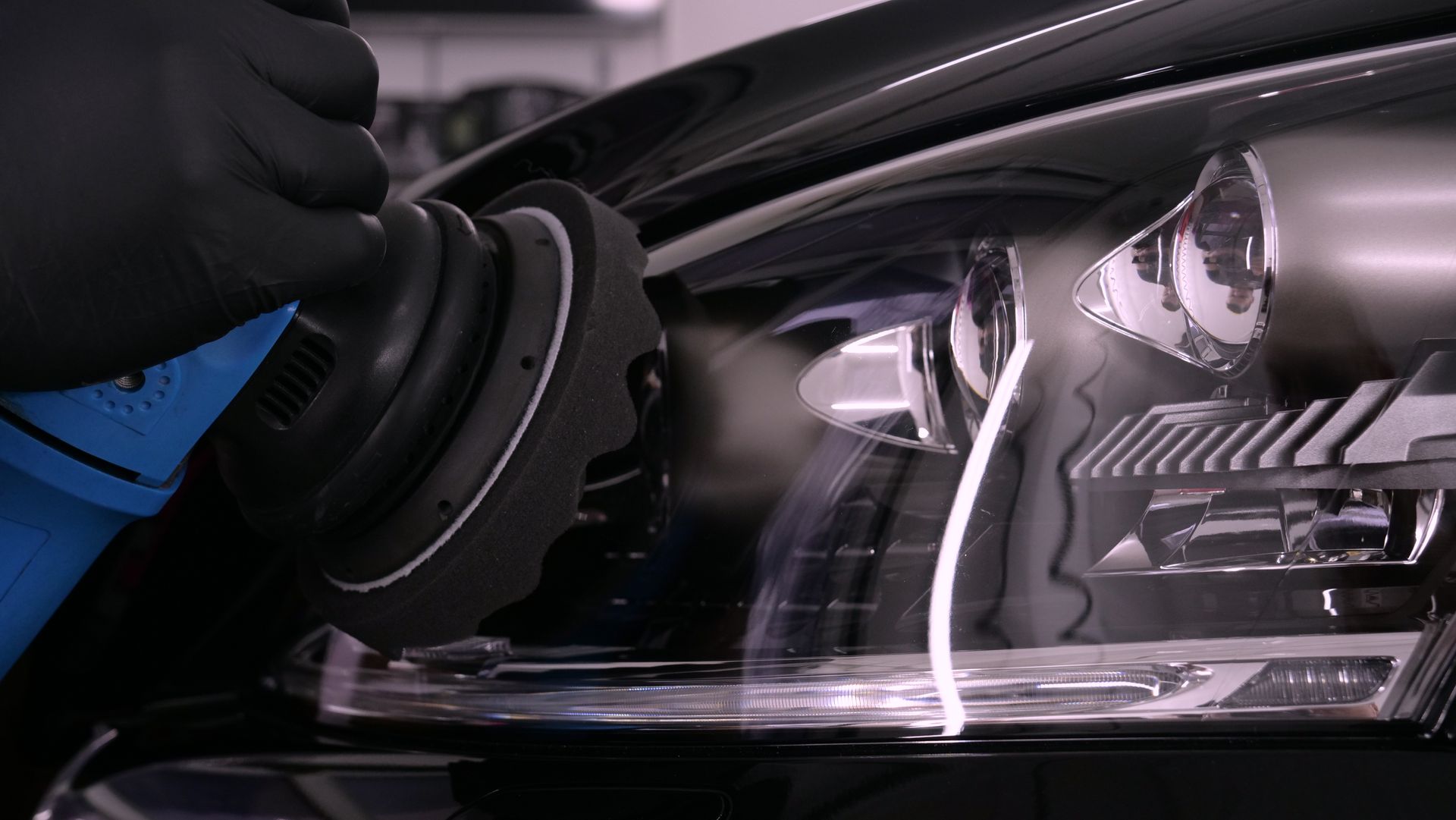 Person in black glove using a polisher on a car headlight, reflecting light, in a garage setting.