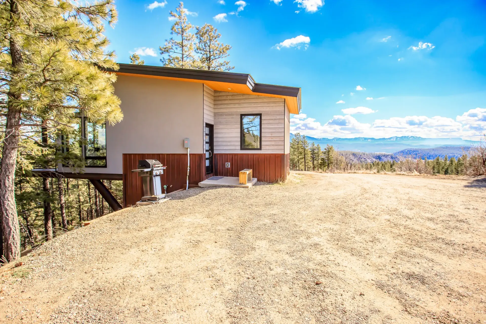 A modern, single-story house with tan and wood siding sits on a dry, dirt hillside under a bright blue sky.
