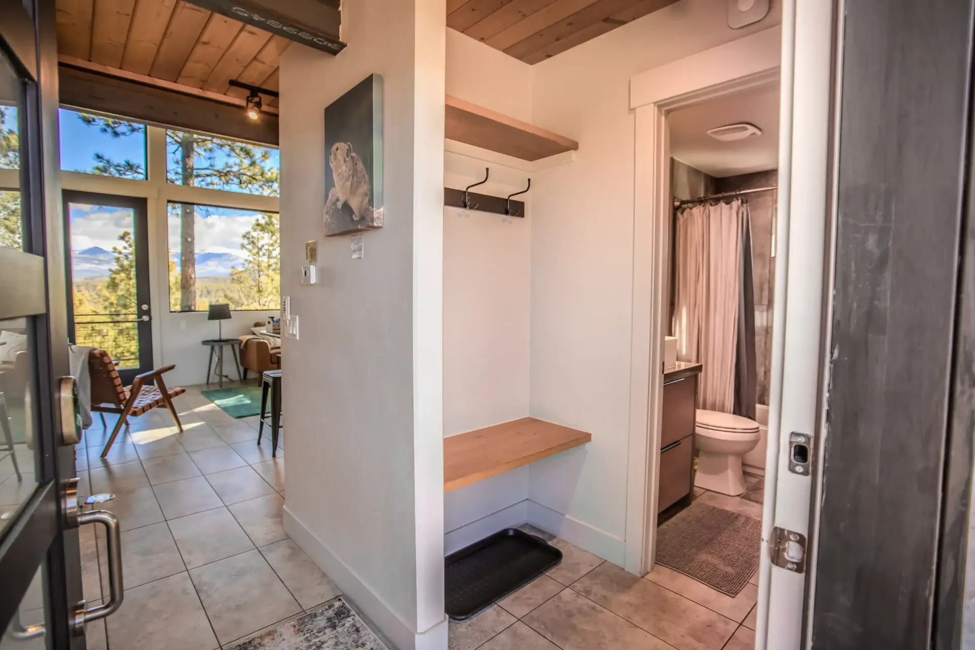 A view from a doorway into a home, showing a mudroom bench and hooks next to a bathroom, with a living area beyond.