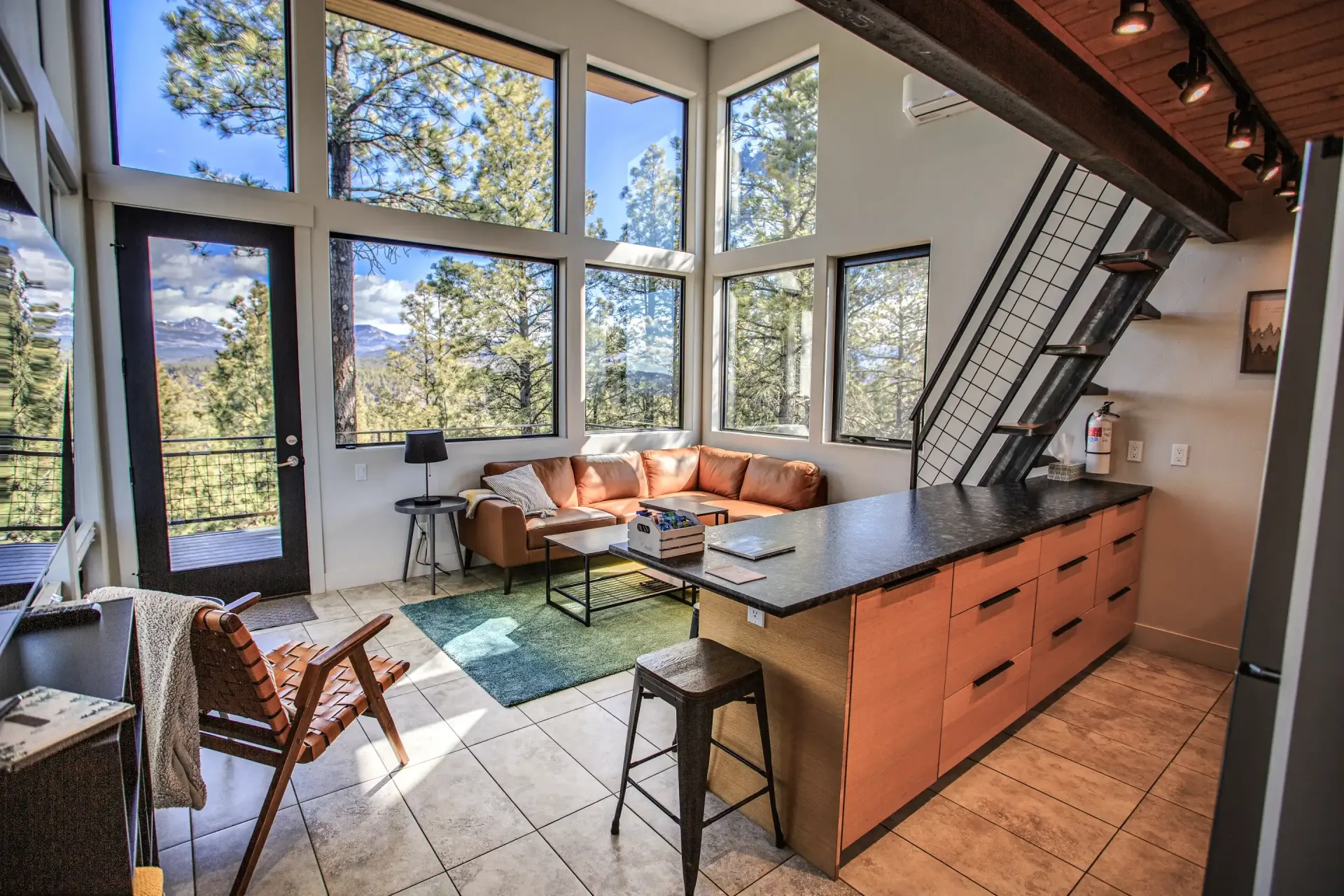 A modern open-plan living room with large windows, a tan leather sofa, a kitchen island with stools, and a metal staircase.