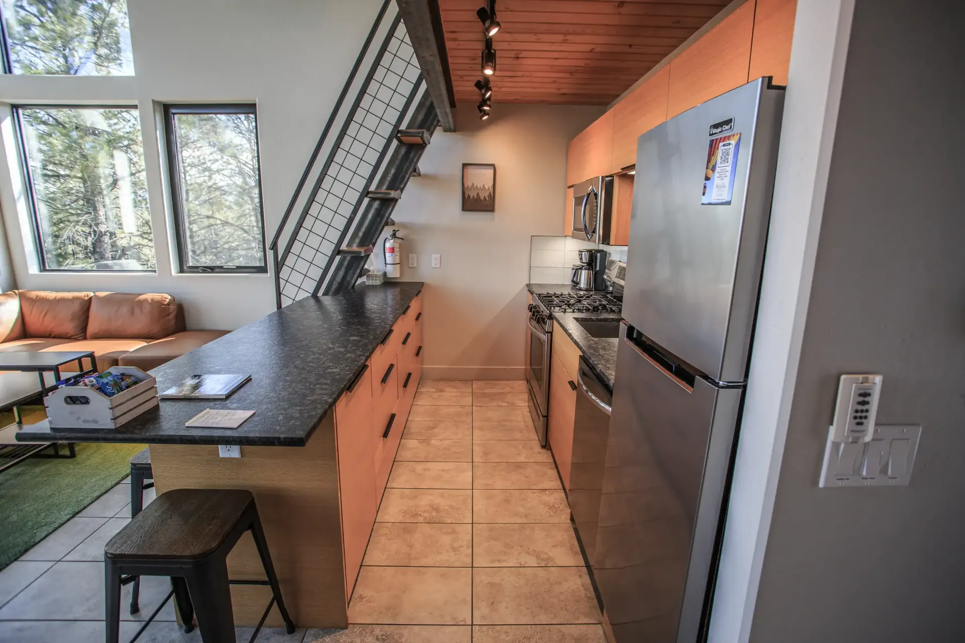 A modern kitchen with light wood cabinets, a stainless steel refrigerator, black countertops, and a tile floor.