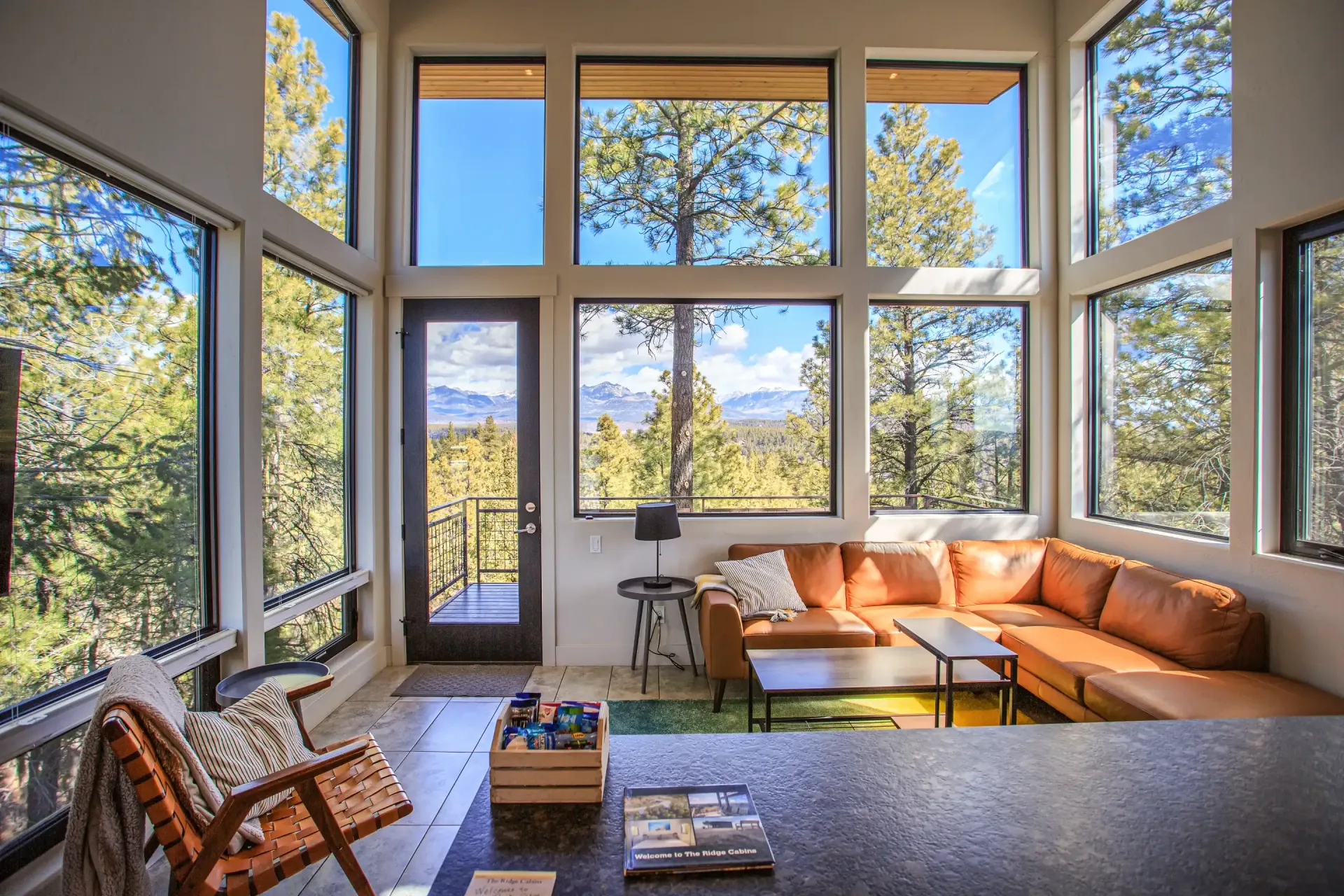 A sun-drenched living room with large windows, a tan sectional sofa, and a wood-framed chair overlooking snowy mountains.