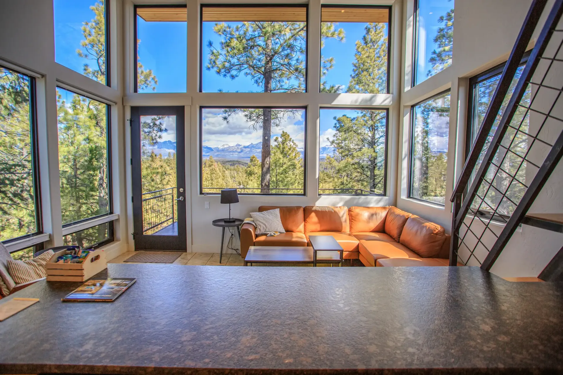 Modern living room with floor-to-ceiling windows showing a forest and mountain view, featuring a tan L-shaped sofa.