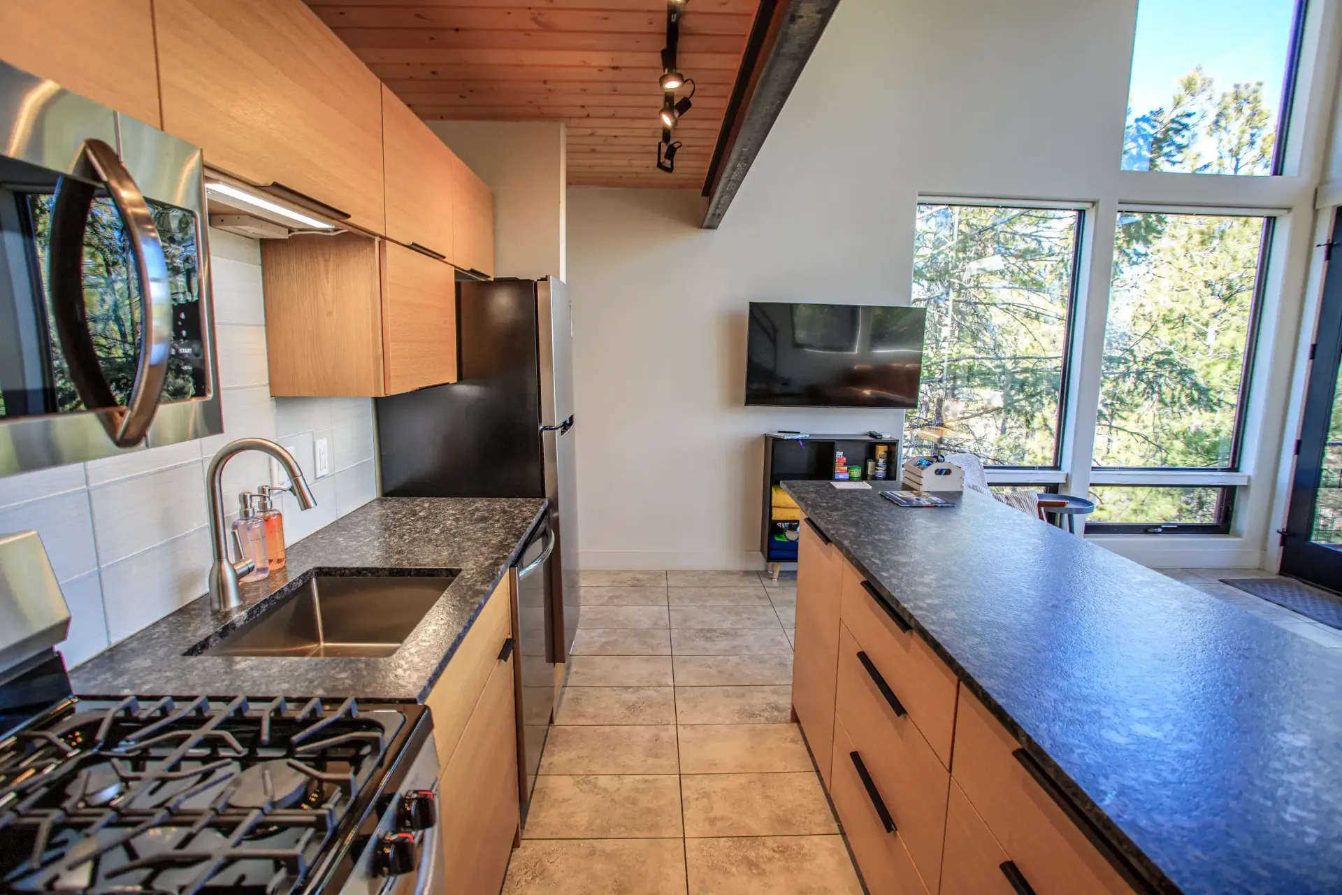 A modern kitchen with light wood cabinets, black countertops, stainless steel appliances, and large windows.