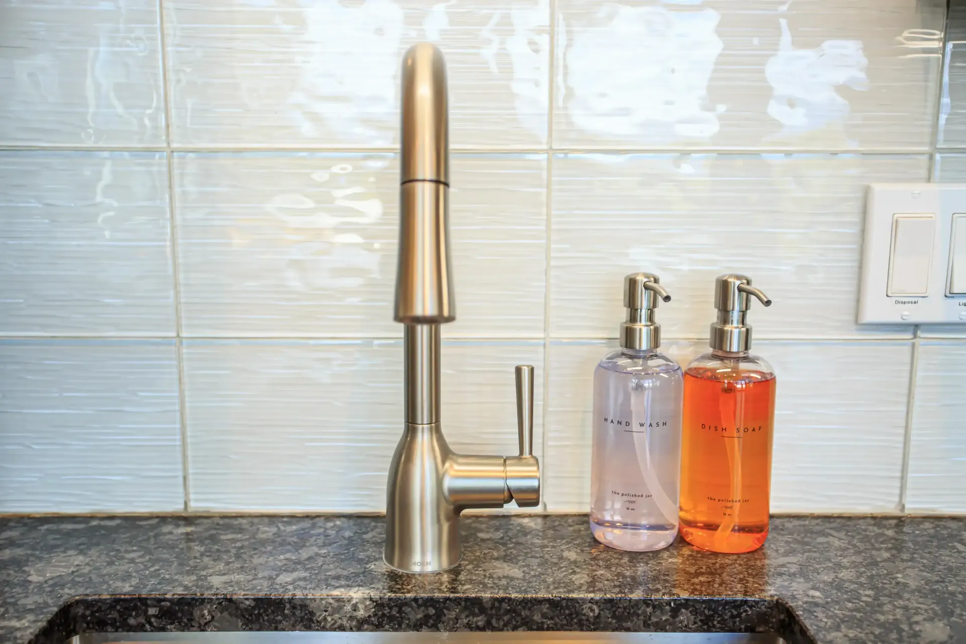 A brushed-nickel pull-down faucet sits on a dark granite counter next to two soap dispensers against white tiled walls.