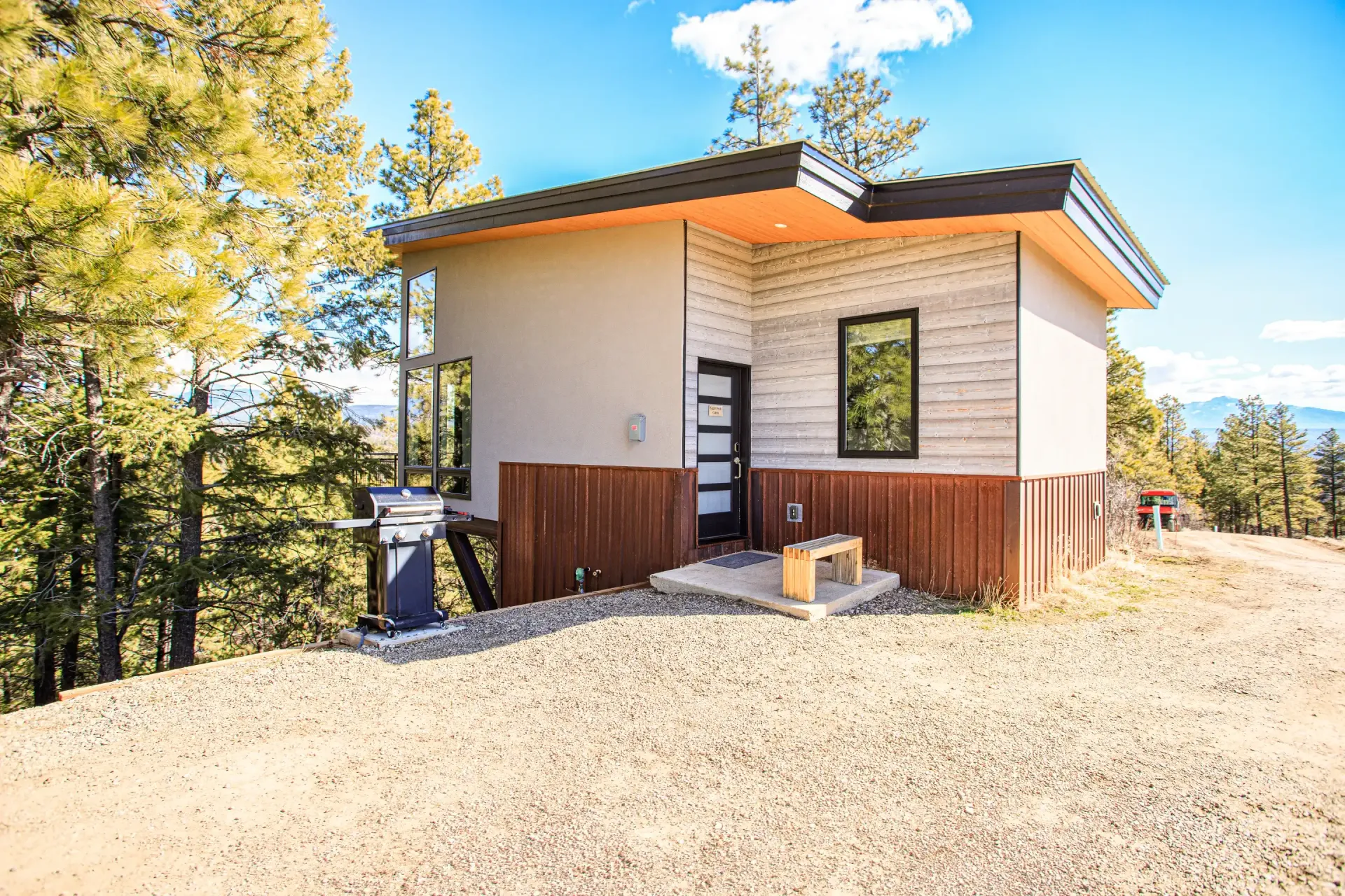 A small, modern cabin with a flat roof and mixed-material siding stands on a gravel lot beside a forest.