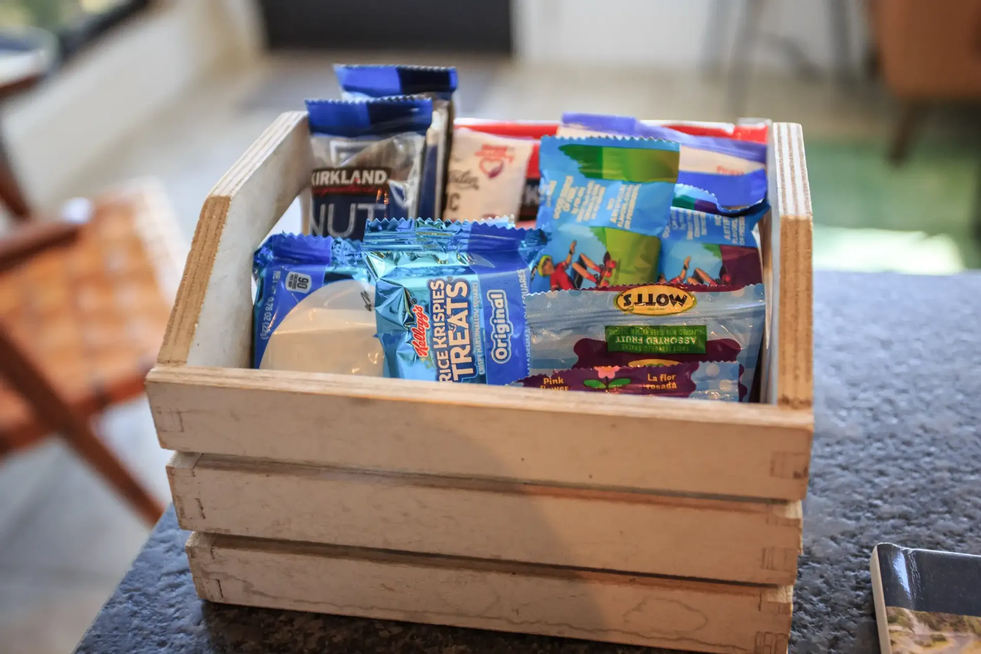 A light wooden crate filled with various snack bags sits on a dark, textured surface.
