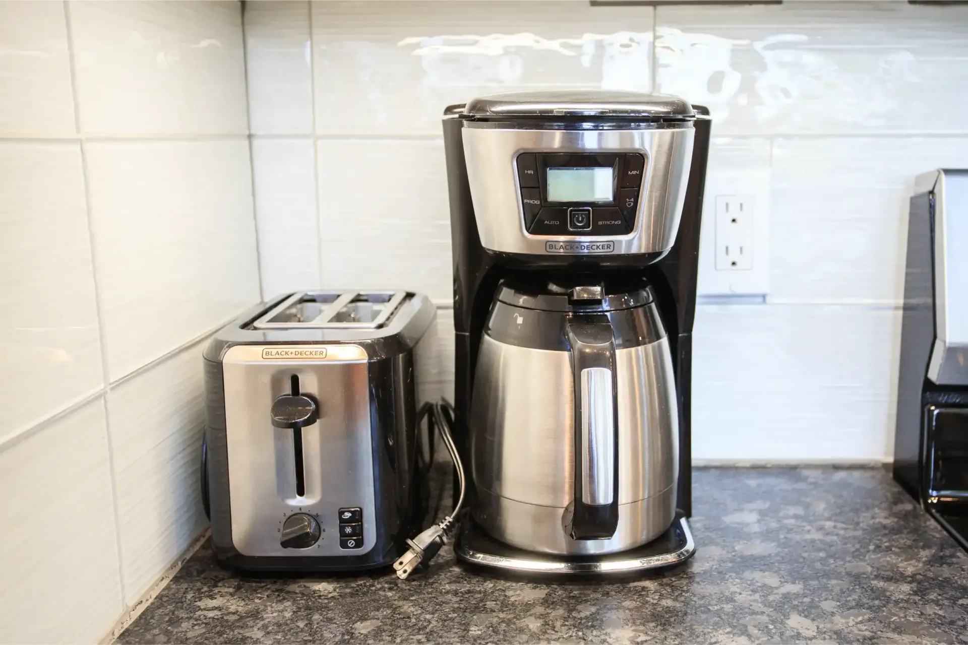 A black and stainless steel toaster and coffee maker sitting on a kitchen counter against a white tiled backsplash.