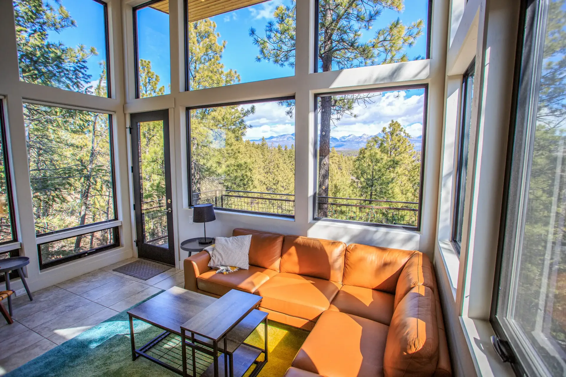 A bright sunroom with large windows overlooking a forest, featuring a tan sectional sofa and two wooden coffee tables.