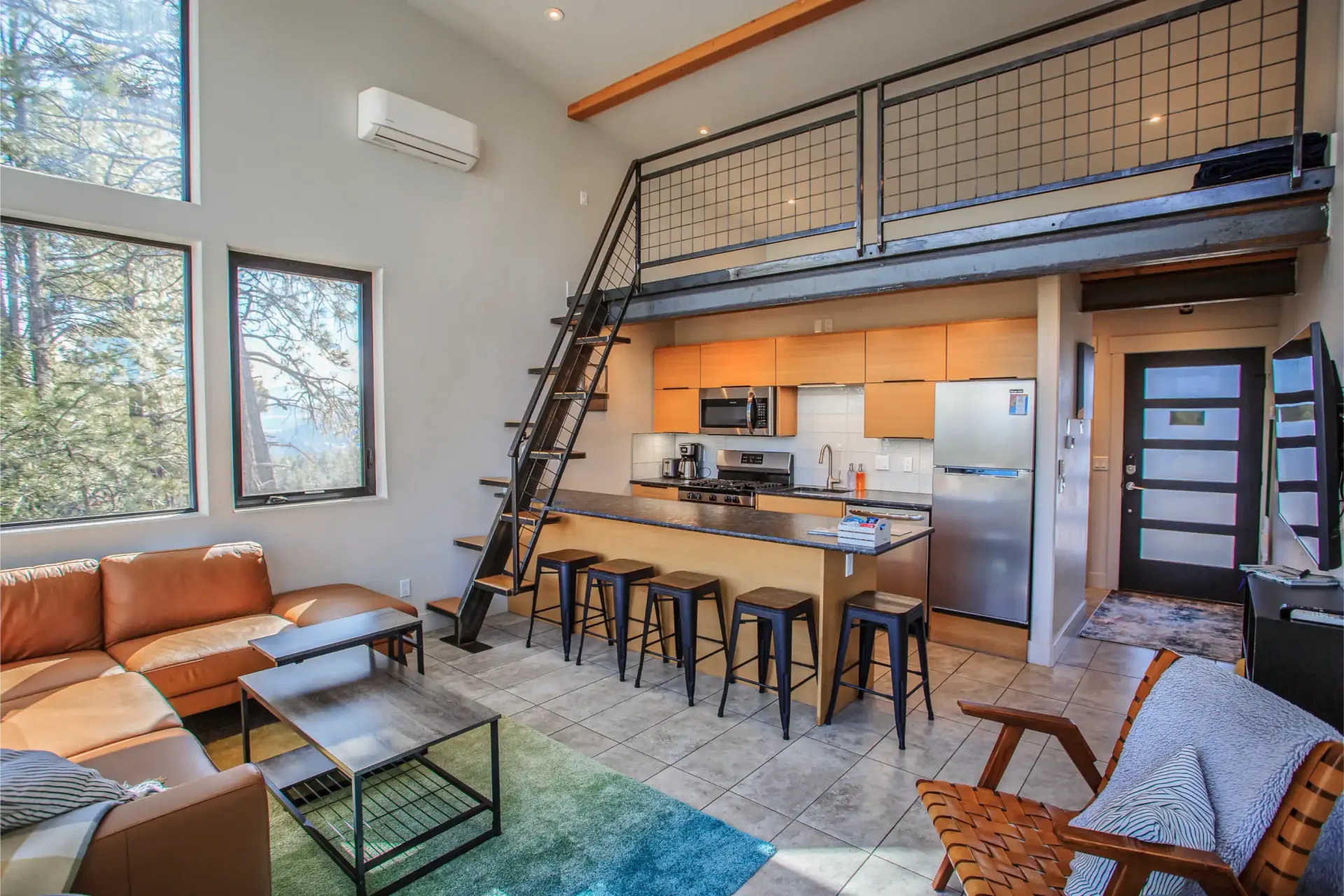 A modern loft-style apartment featuring an open kitchen, a brown leather sofa, a metal ladder, and a mezzanine level.