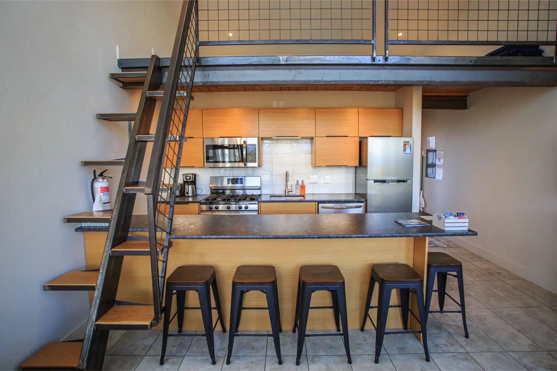 A kitchen with light wood cabinets, stainless steel appliances, a granite island with four stools, and a metal loft ladder.