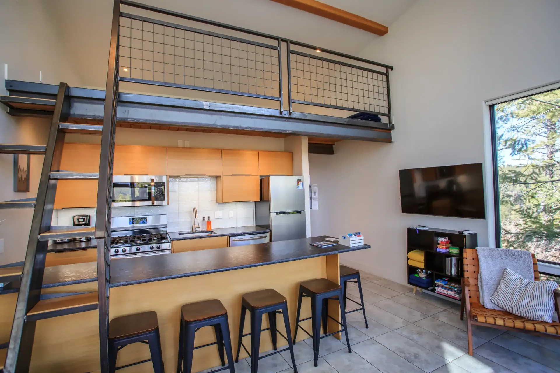 A kitchen with a lofted level, featuring a long counter with four black stools, stainless appliances, and a TV area.
