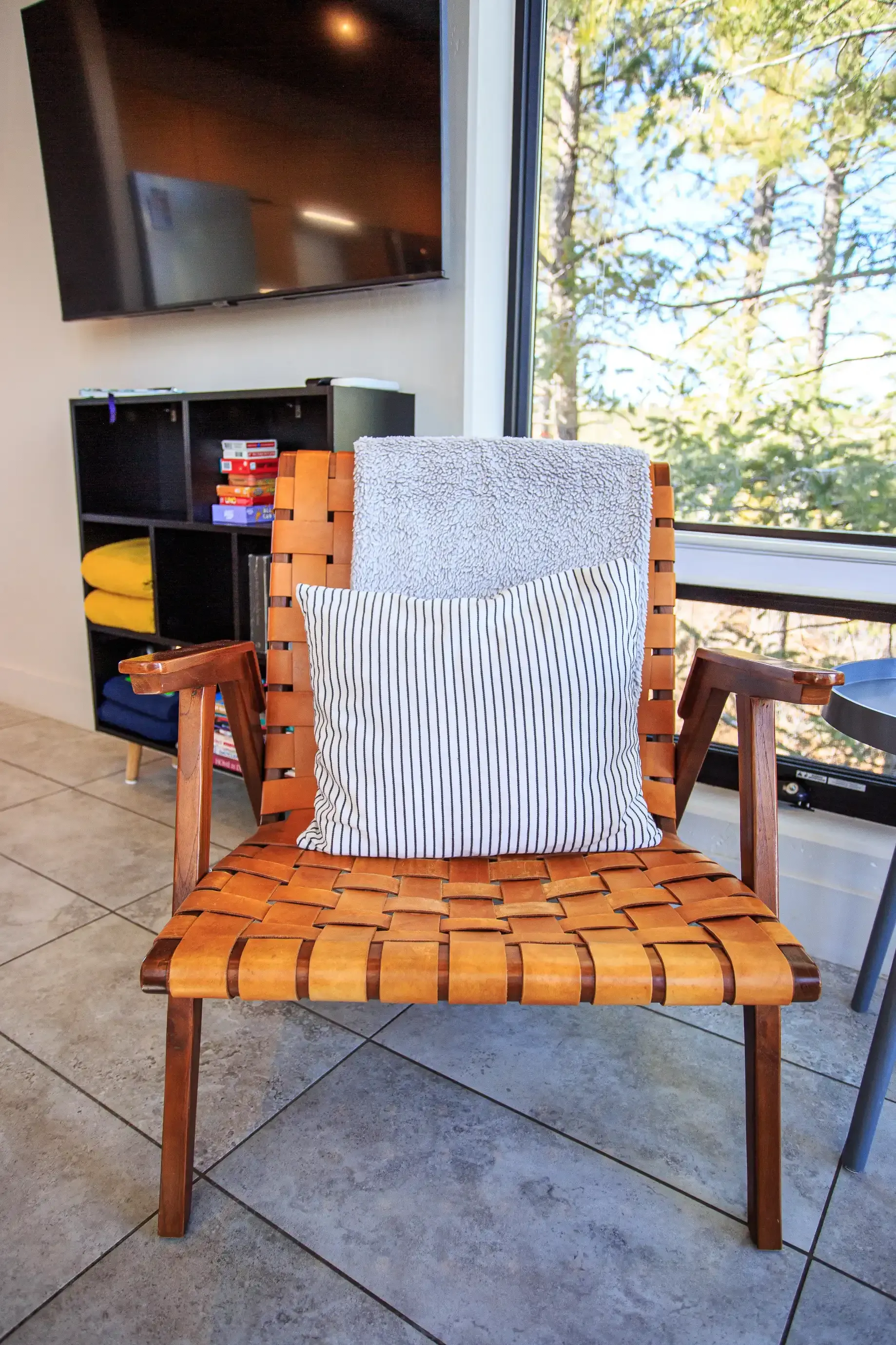 A leather strap lounge chair with a patterned throw pillow, positioned near a window and a bookshelf.