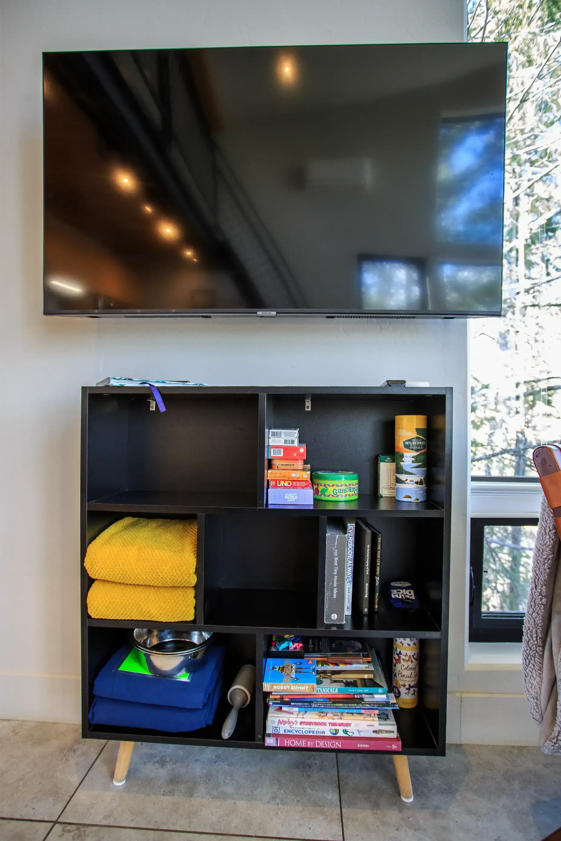A wall-mounted TV sits above a black, tiered storage shelf filled with folded towels, books, and miscellaneous items.