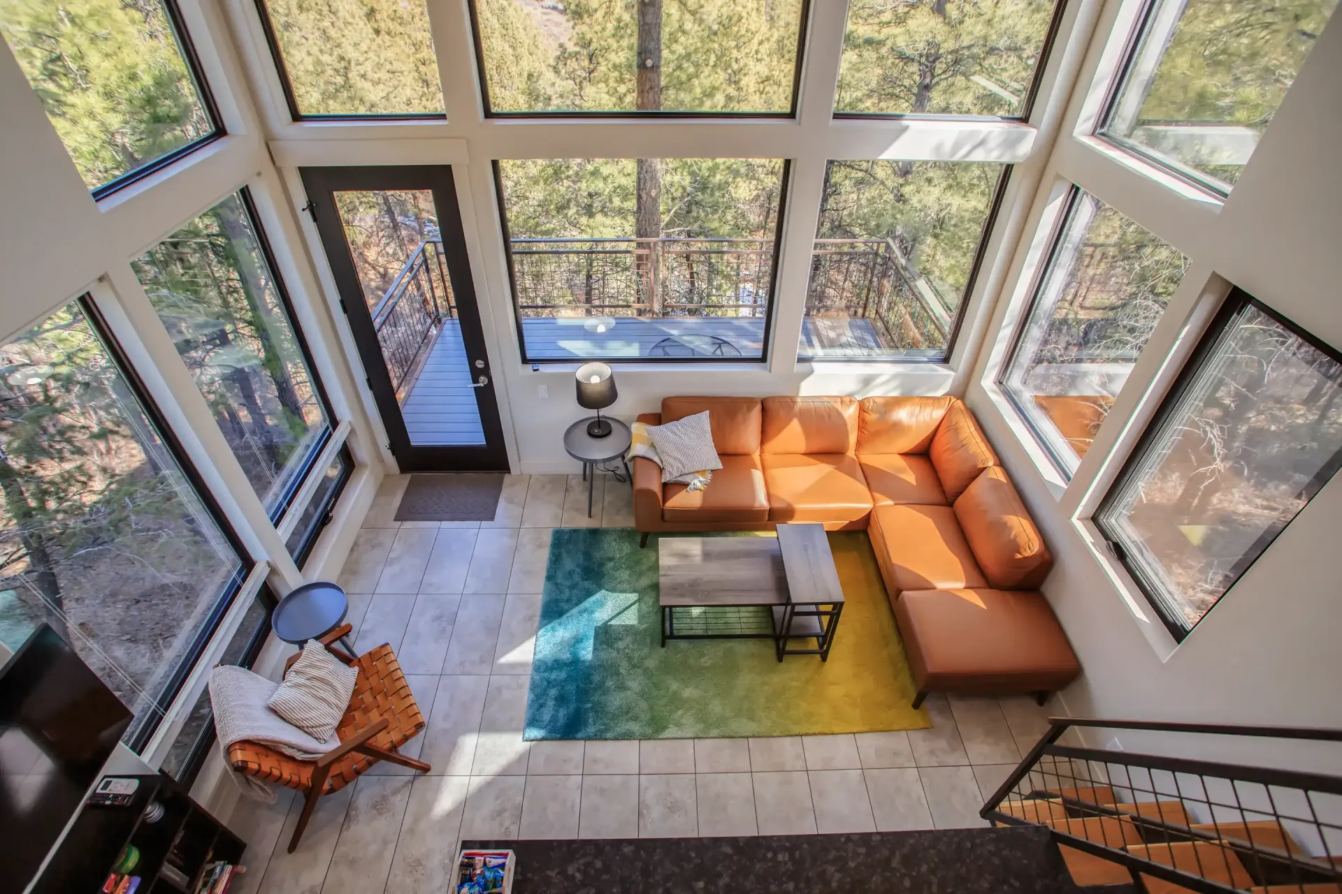 A high-angle view of a sunlit living room with large windows, a tan sectional sofa, a colorful rug, and an armchair.
