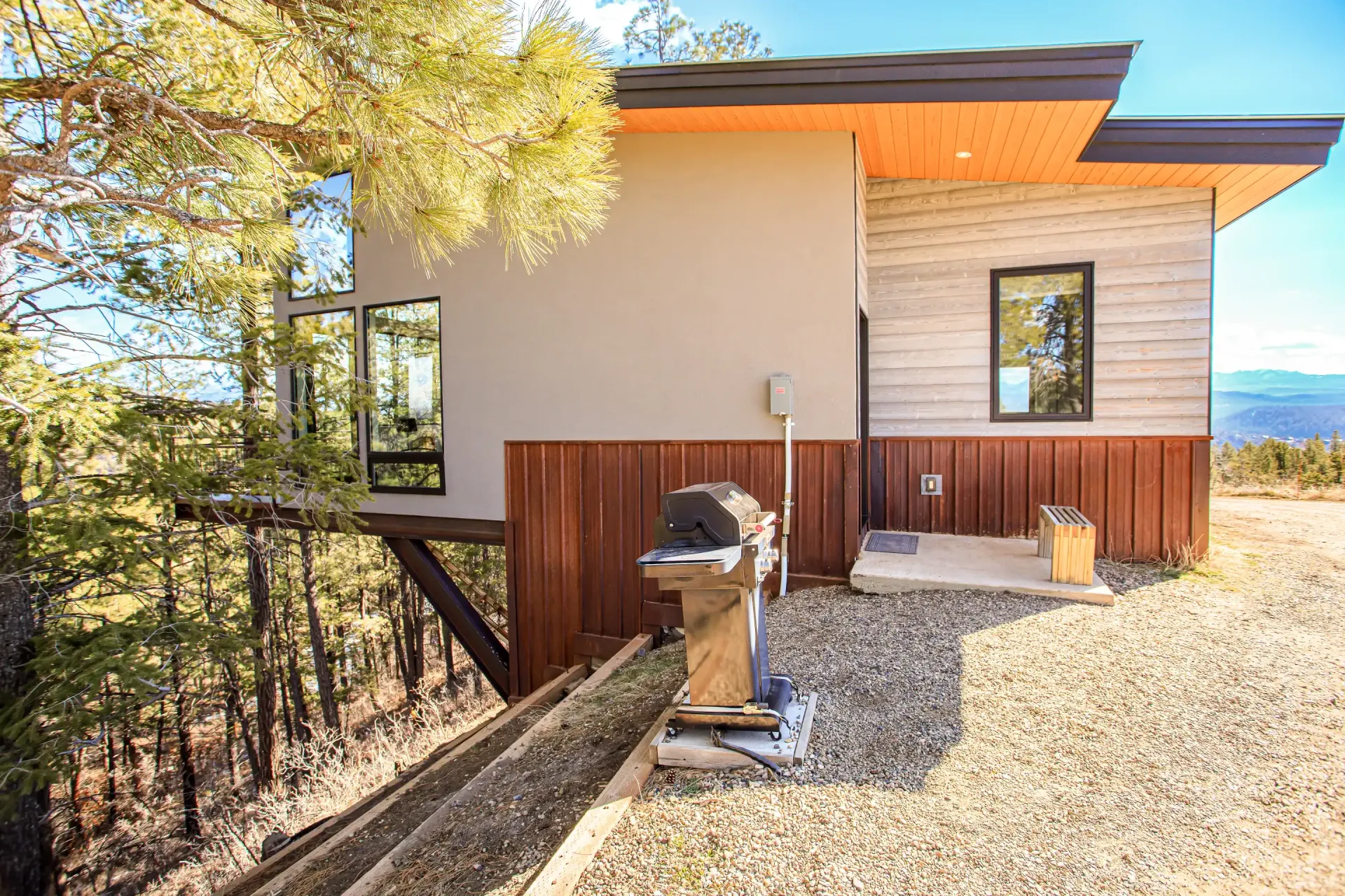 A modern hillside house with beige walls, wood paneling, and a grill on a gravel patio under a clear blue sky.