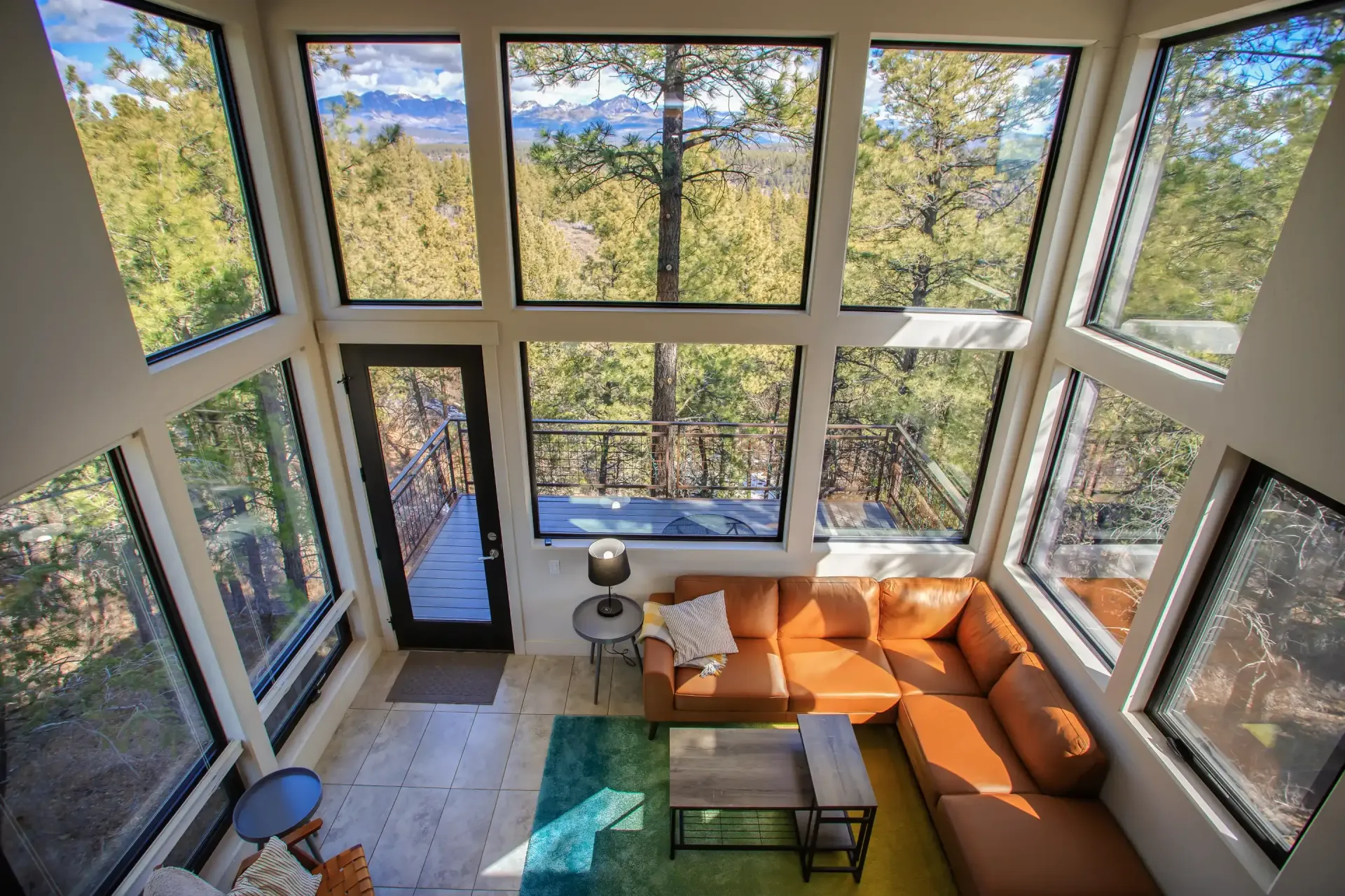 A living room with a brown leather sectional, floor-to-ceiling windows, and a scenic forest view from an elevated deck.