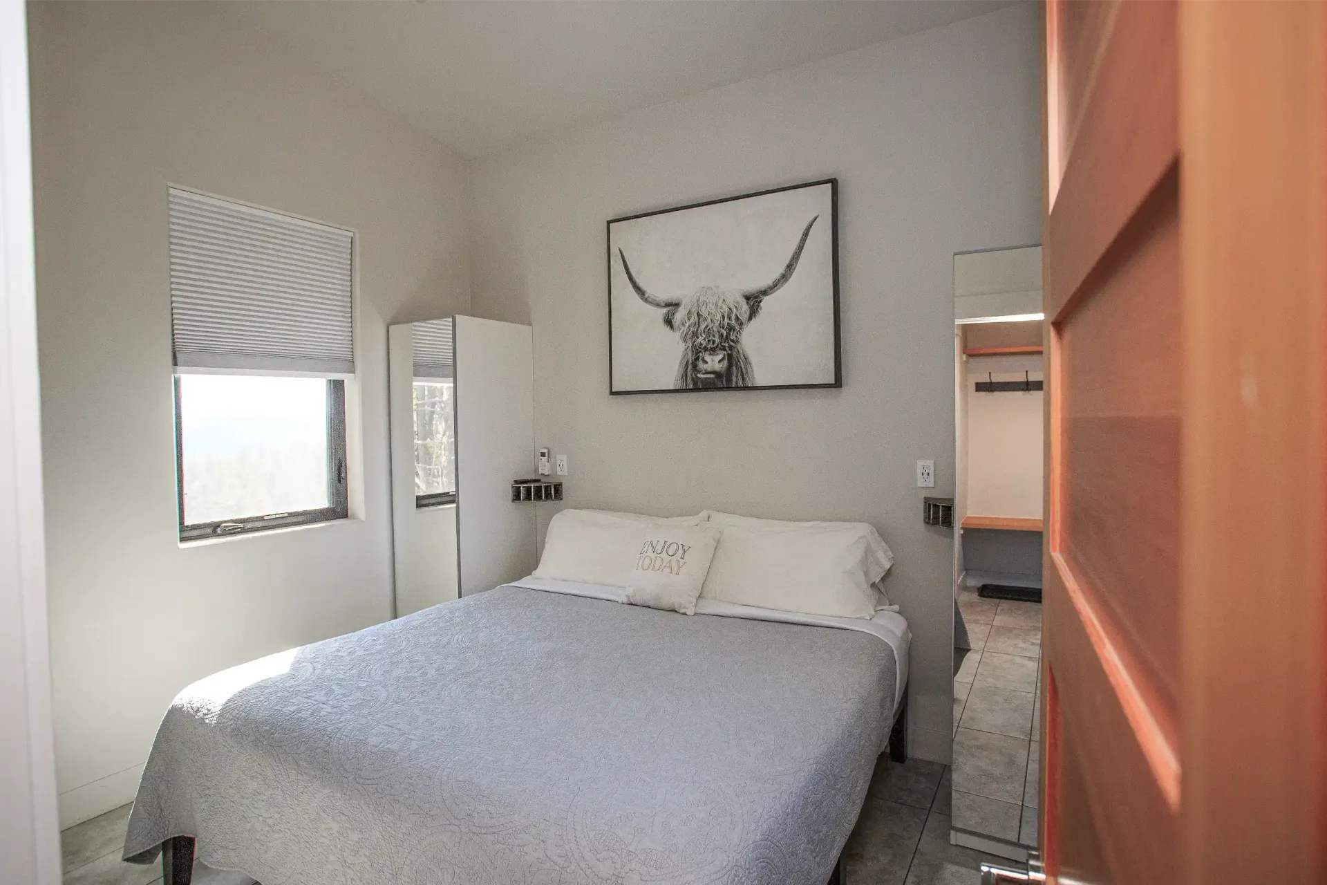 A small, neutral-toned bedroom with a bed, white bedding, a Highland cow wall print, and a mirror next to the door.