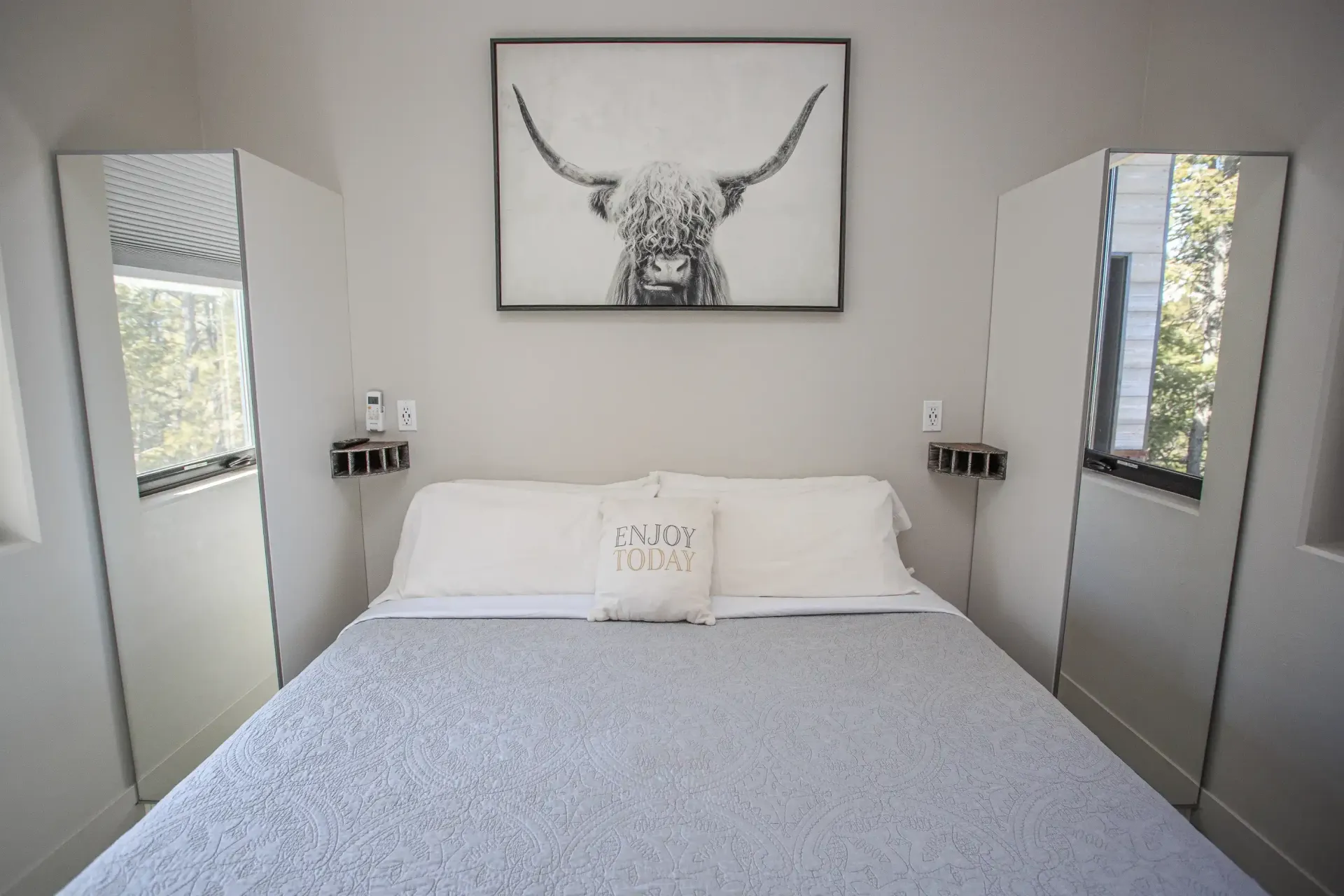 A bed with white bedding in a neutral-toned room, featuring a framed Highland cow print above and side mirrors.