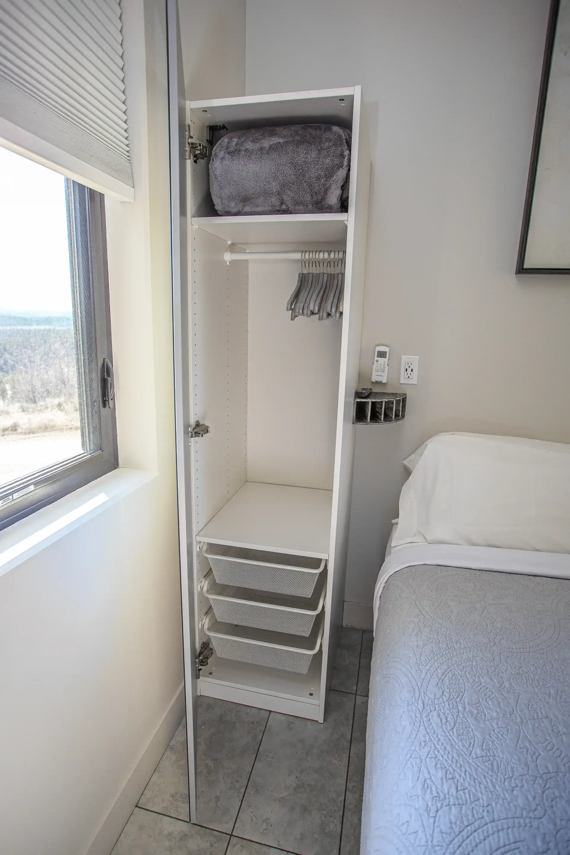 A white, narrow, open wardrobe stands beside a bed, featuring a top shelf with a patterned item and three wire baskets.